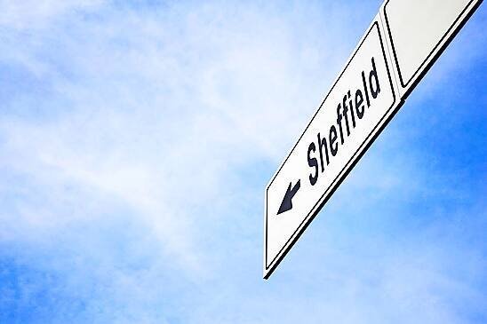 White signboard with an arrow pointing left towards Sheffield, England, United Kingdom, against a hazy blue sky in a concept of travel, navigation and direction